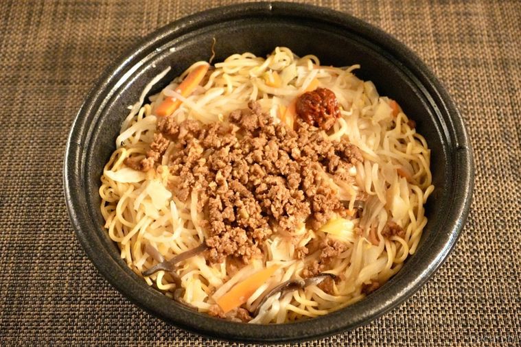 茶色いランチョンマットにのったコンビニの豚そぼろのせにんにく焼きそばが入った黒い容器