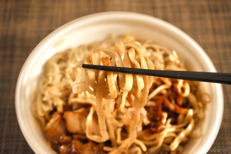 茶色いランチョンマットにのった白い容器のコンビニの旨辛二郎系ラーメンの麺を黒い箸で持ち上げる様子