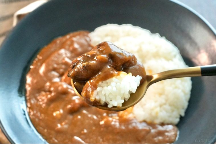木のカッティングボードと黒い器にのったレトルトカレーを金色と黒色のスプーンですくう様子