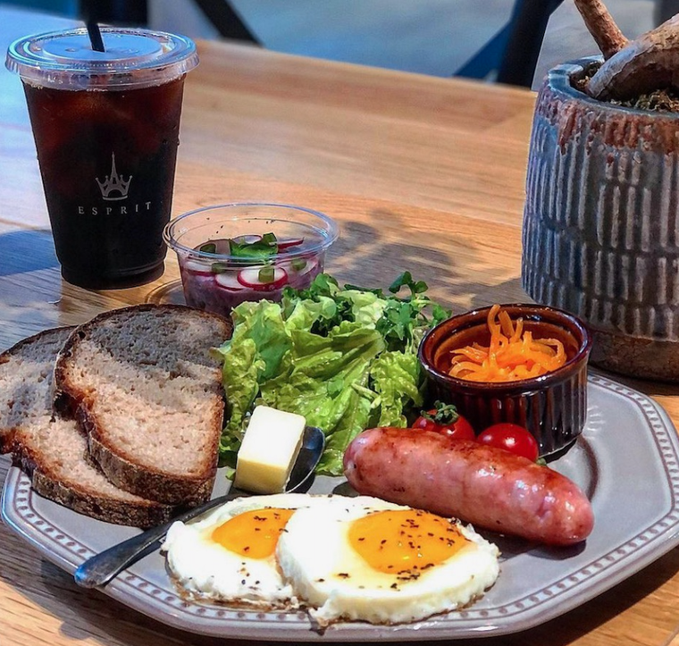 グレーのお皿に卵、ソーセージ、ライ麦パン、サラダ、にんじんがのるプレート。アイスコーヒー