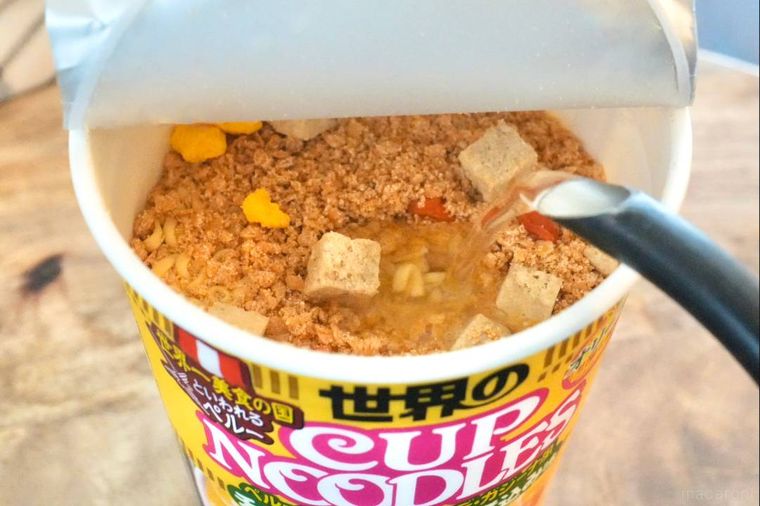木のカッティングボードにのった黄色いパッケージのカップヌードルに熱湯を注ぐ様子