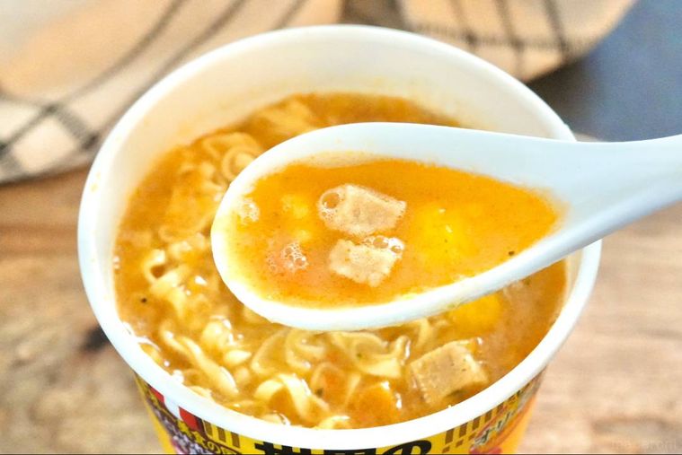 木のカッティングボードにのったカップヌードルのスープを白いレンゲで飲む様子