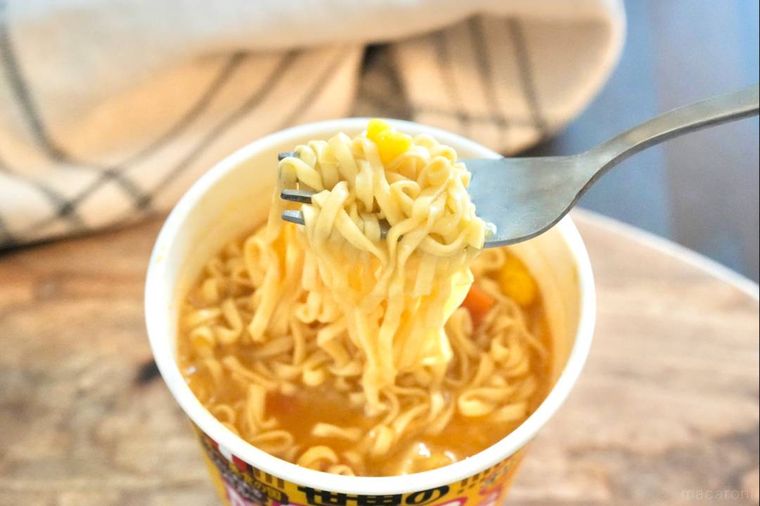 木のカッティングボードにのったカップヌードルを銀のフォークで食べる様子