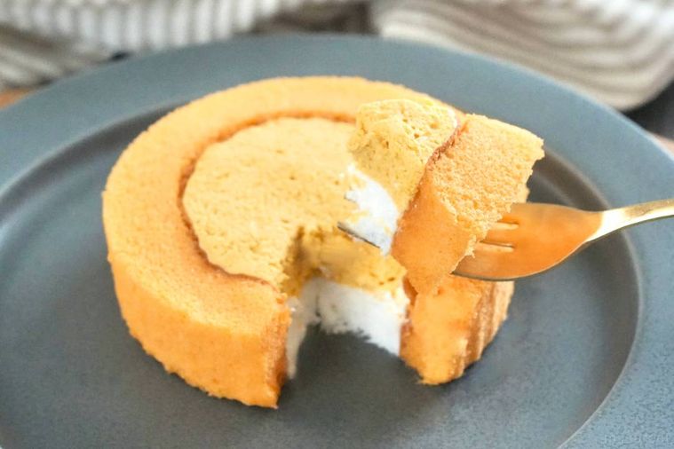 木のカッティングボードとグレーの皿にのったかぼちゃロールケーキを金のフォークで食べる様子