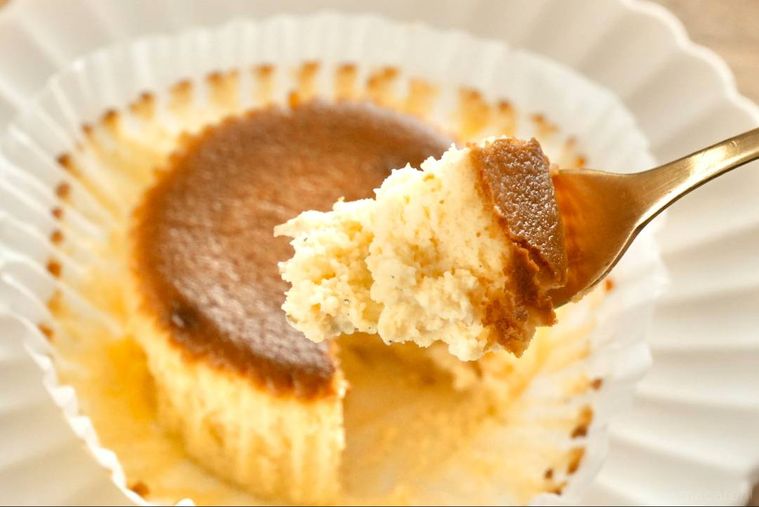 木のカッティングボードと白い皿にのったベイクドチーズケーキを金のフォークで食べる様子