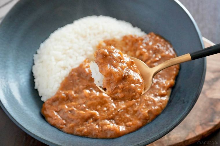 木のカッティングボードにのった黒い器に盛られたキーマカレーと黒色と金色のスプーンで食べる様子
