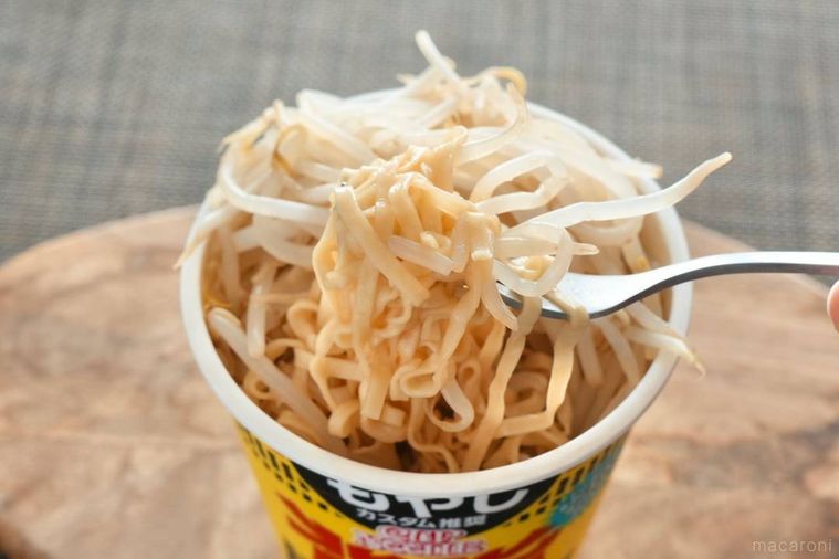 黄色いパッケージのカップヌードルビッグにトッピングしたもやしを銀のフォークで食べる様子