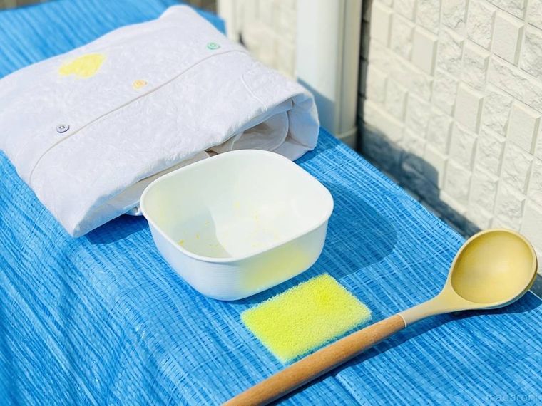 カレーで黄色く染まったお玉、保存容器、食器用スポンジ、白いシャツを日光に当てている様子