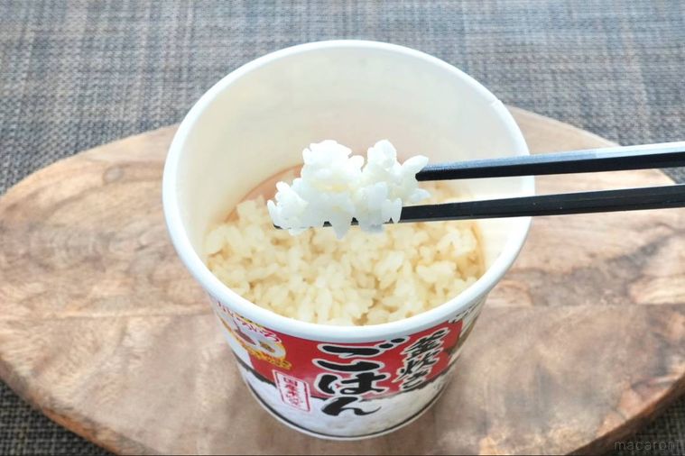 木のカッティングボードにのったカップタイプのインスタント白ごはんを黒い箸で持ち上げる様子