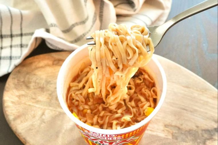 木のカッティングボードにのった赤いパッケージのスパイスカレーヌードルを銀のフォークで持ち上げる様子