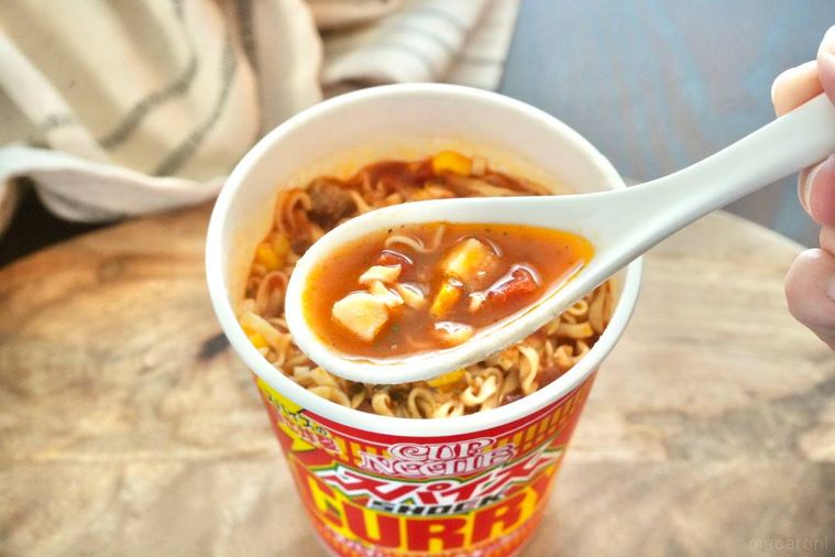 木のカッティングボードにのった赤いパッケージのスパイスカレーヌードルのスープを白のれんげですくう様子