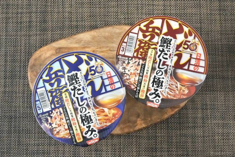 木のカッティングボードにのった鰹だしのカップうどんどん兵衛の赤色と青色のパッケージ
