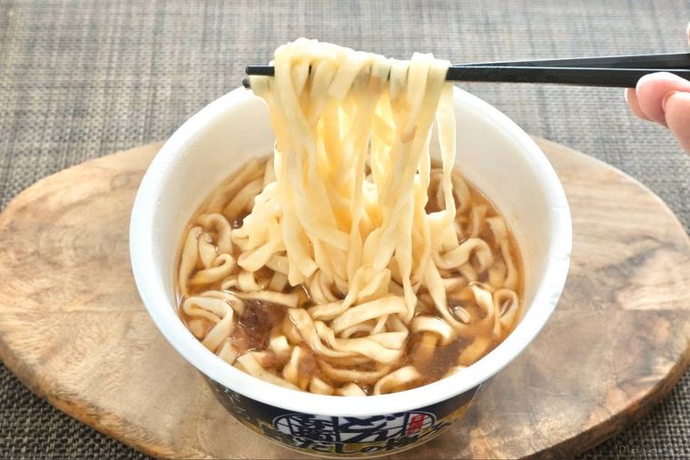 木のカッティングボードにのったカップうどんを黒い箸で持ち上げる様子