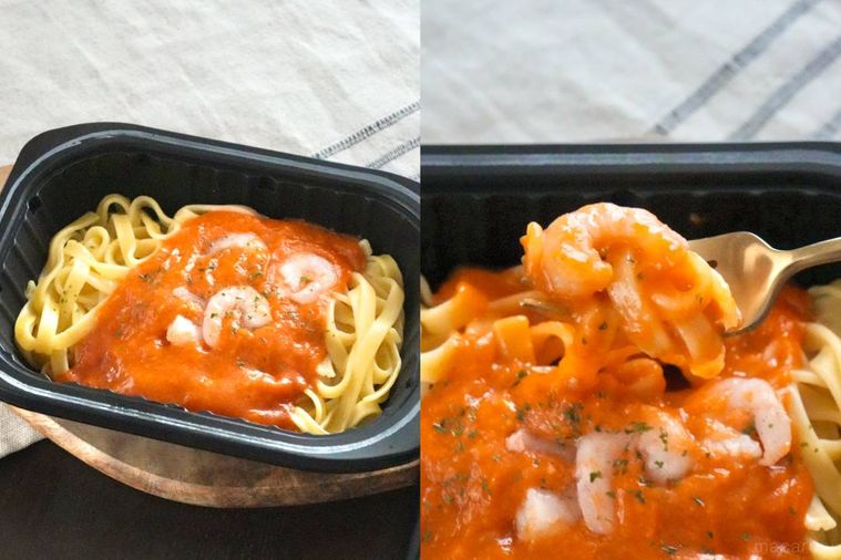 木のカッティングボードにのった黒いトレー入りの冷凍生パスタ海老トマトクリームを金のフォークで食べる様子