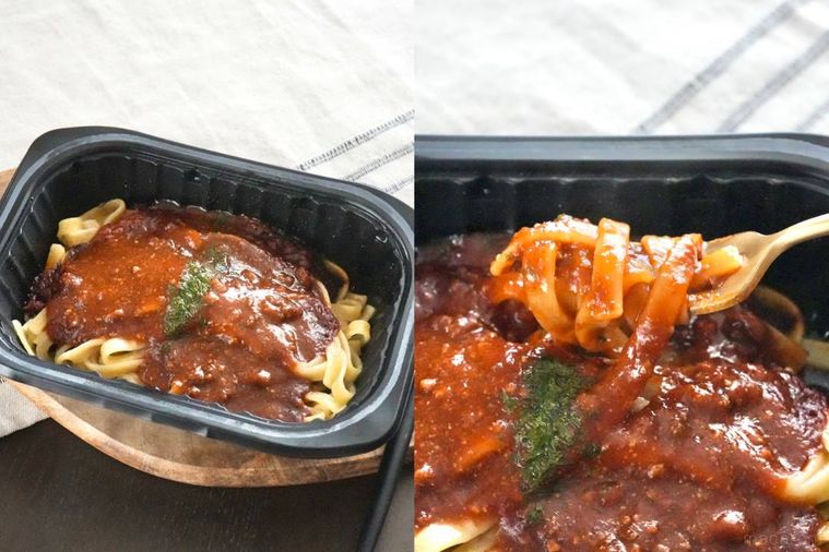 木のカッティングボードにのった黒いトレー入りの冷凍生パスタボロネーゼを金のフォークで食べる様子