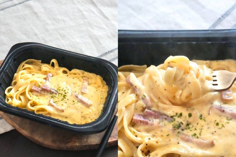 木のカッティングボードにのった黒いトレー入りの冷凍生パスタカルボナーラを金のフォークで食べる様子