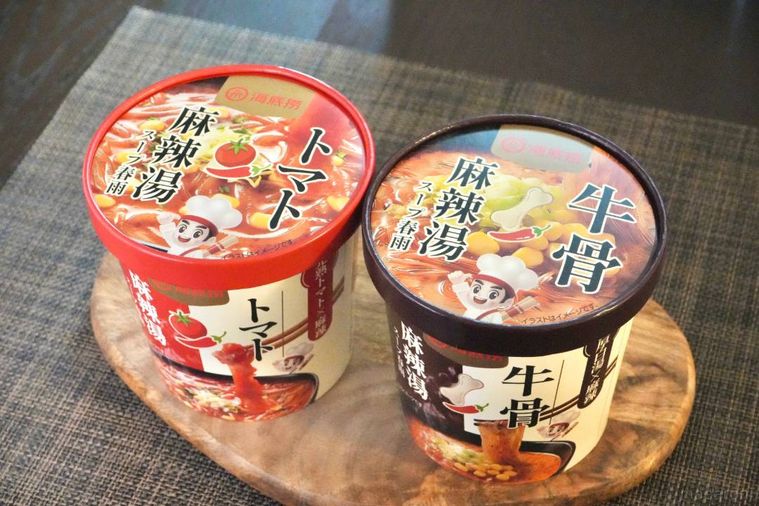 木のカッティングボードにのったトマト味と牛骨スープのカップ麻辣湯の赤色と黒色パッケージ