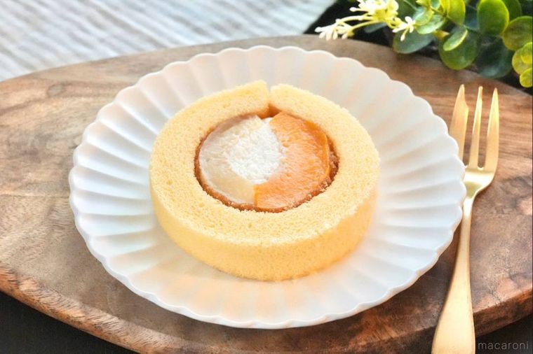 木のカッティングボードと白い皿にのった黄桃と白桃のプレミアムロールケーキと金色のフォーク