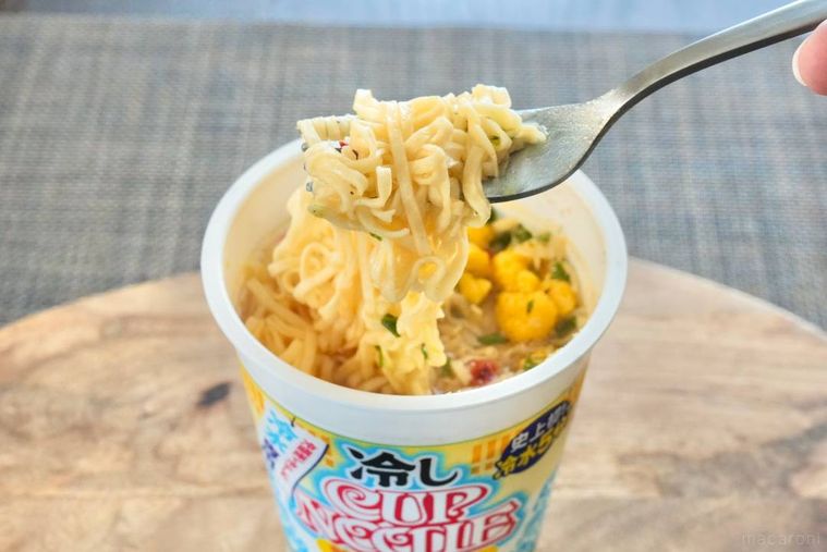 木のカッティングボードにのった冷しカップヌードル鶏塩レモン味の麺を銀色のフォークで持ち上げる様子