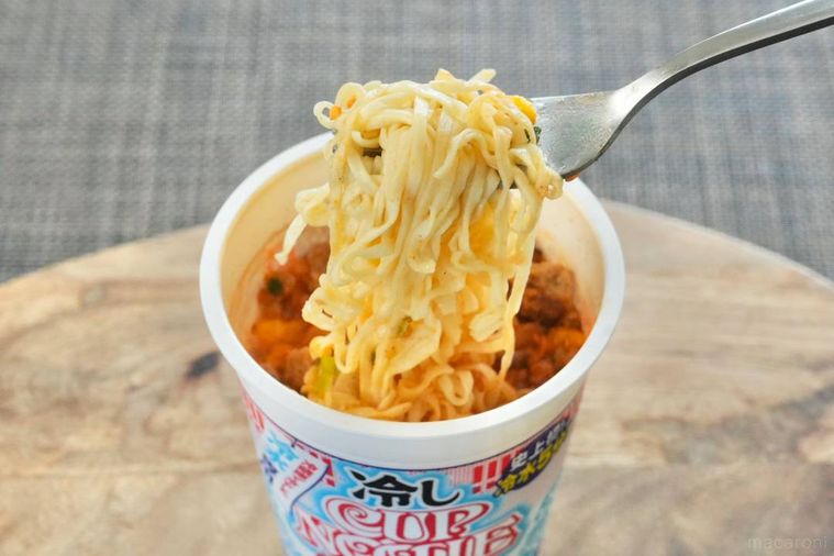 木のカッティングボードにのった冷しカップヌードルピリ辛キムチ味の麺を銀色のフォークで食べる様子