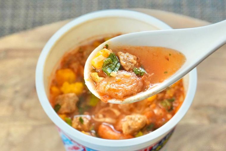 木のカッティングボードにのった冷しカップヌードルピリ辛キムチ味のスープを白いれんげですくう様子