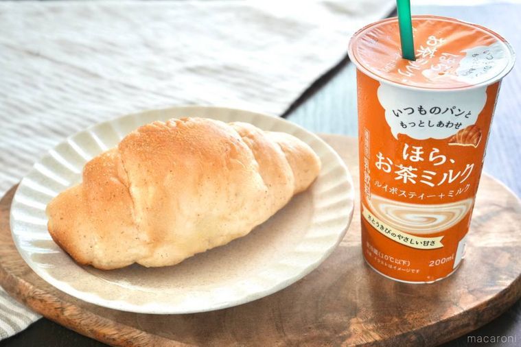 木のカッティングボードにのったオレンジ色のルイボスティーミルクのカップ飲料と白い器に盛られた塩パンとストライプの布