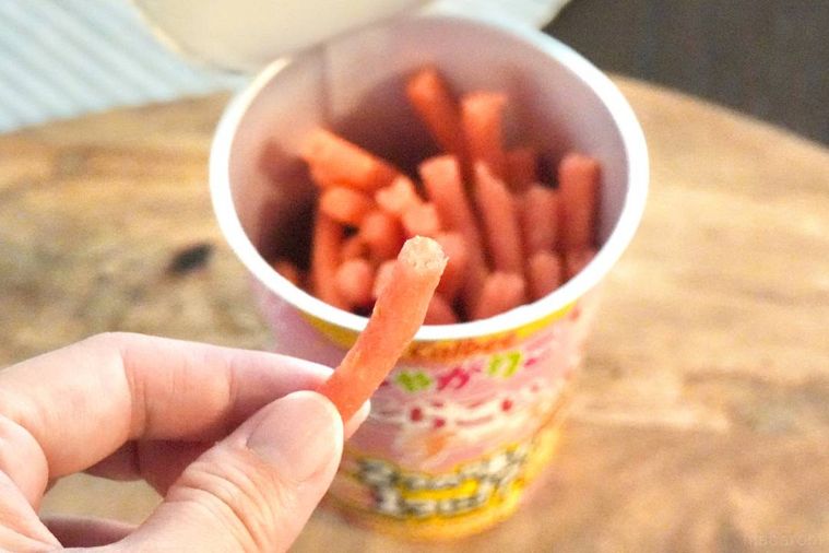 木のカッティングボードにのった黄色とピンク色のじゃがりこのパッケージと中身を指でつまみ上げる様子