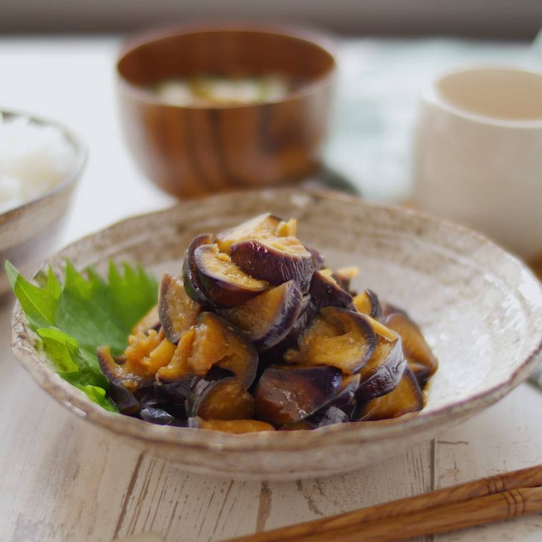 なすの甘酢しょうゆ漬けを盛り付けた薄茶色の小皿に、ご飯と味噌汁が添えられている様子