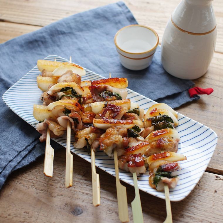 焚き火で炭火焼きされた旨辛長芋と豚バラ肉の串焼きが香ばしく焼かれて長方形の和風の器に盛り付けられているところ