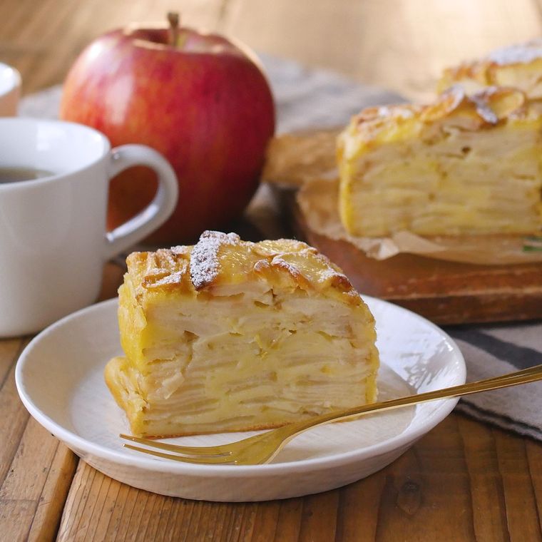 切って白い皿に盛り付けたガトーインビジブルとフォークに、コーヒー入りの白いマグカップとまるごとのりんごが添えられている