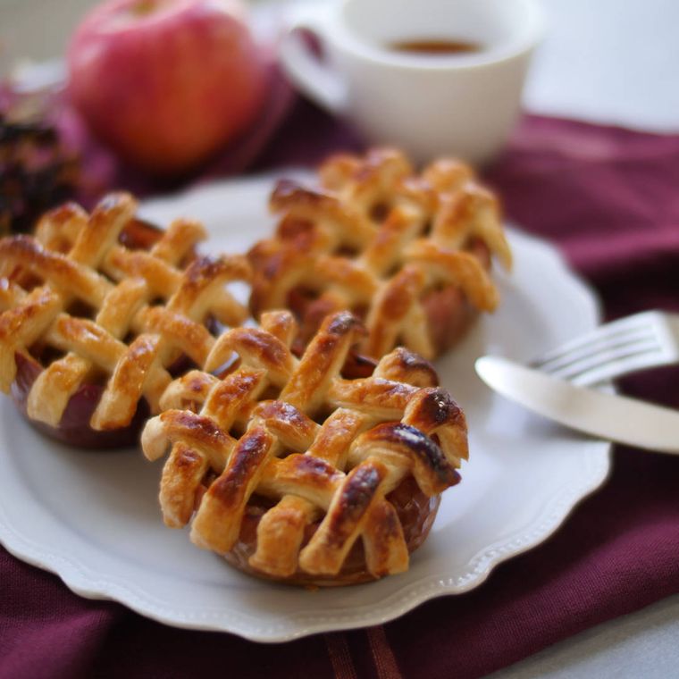 白い皿に盛られたまるごとりんごパイ3個にフォークとナイフが立てかけられており、紅茶入りのマグカップとりんごが奥にある