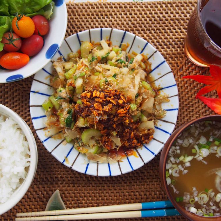 縞模様の白い皿に盛られた食べるラー油で無限セロリに、ごはん・みそ汁・麦茶・ミニトマトのサラダが添えられている様子