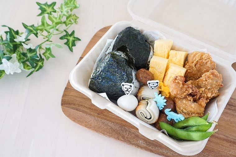 おにぎりとおかずを盛り付けたバガスランチボックスを木の板にのせ、緑の葉と共にテーブルに並べた様子