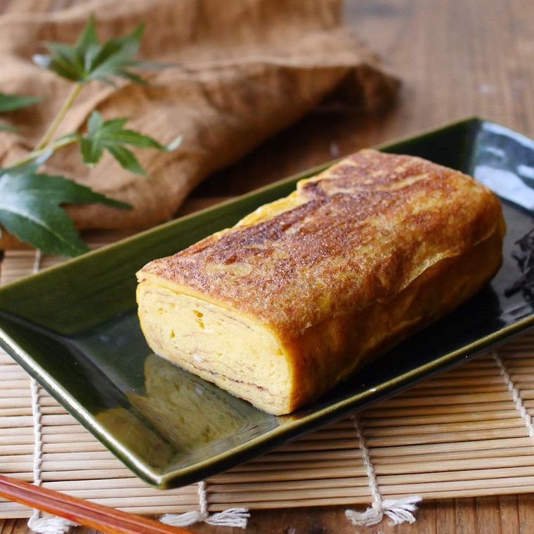 濃色の長皿に盛りつけられている厚焼き卵、巻きすや箸など