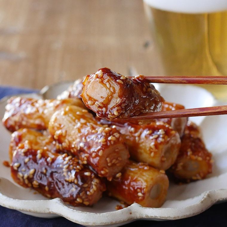 花びら形の白い皿に盛りつけられている餅入りちくわのピリ辛炒めを箸で持ち上げており、奥にビール入りのグラスが添えられている