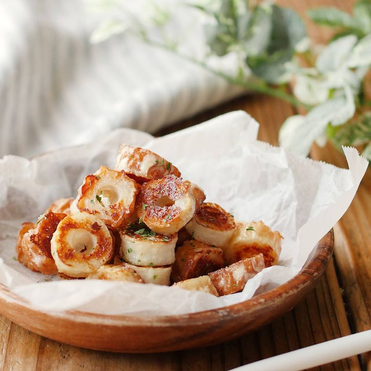 木製皿の上の敷き紙に盛りつけられたちくわのイタリアン風ソテーの奥に、ストライプ模様のクロスが折りたたまれて添えられている