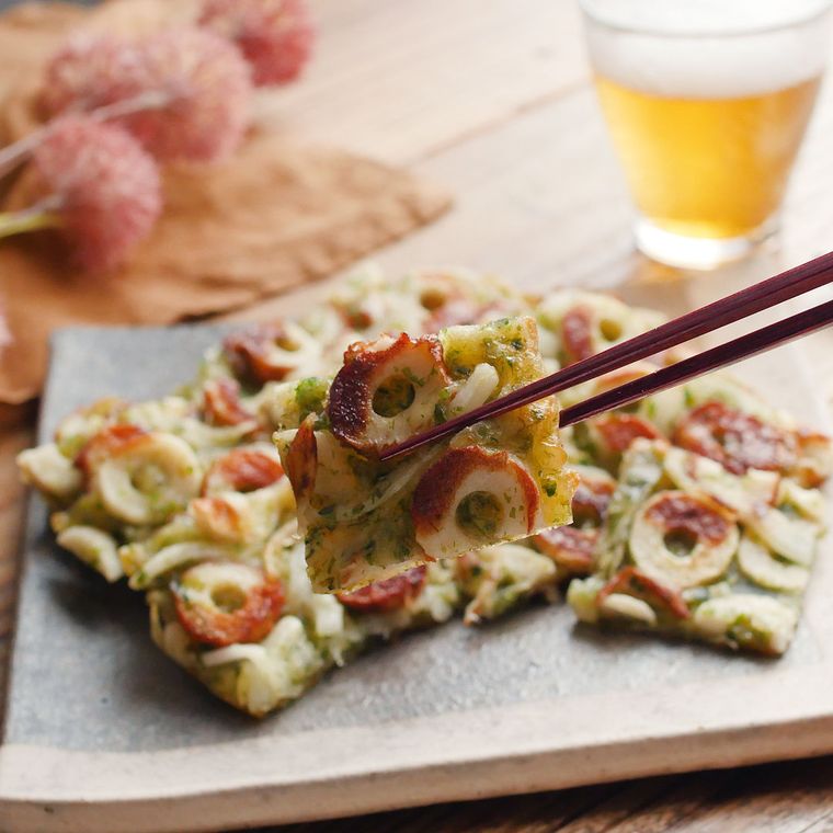 角皿に盛りつけられたちくわの磯辺チヂミ風を箸でつまんでいる様子と、奥に置かれたビール入りのグラス