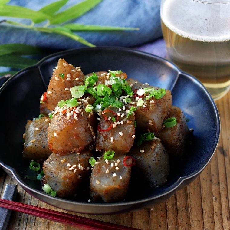 黒い器に盛られたこんにゃくの旨辛炒めに、箸置きに置かれた箸・ビール入りのグラス・青いテーブルクロスが添えられている