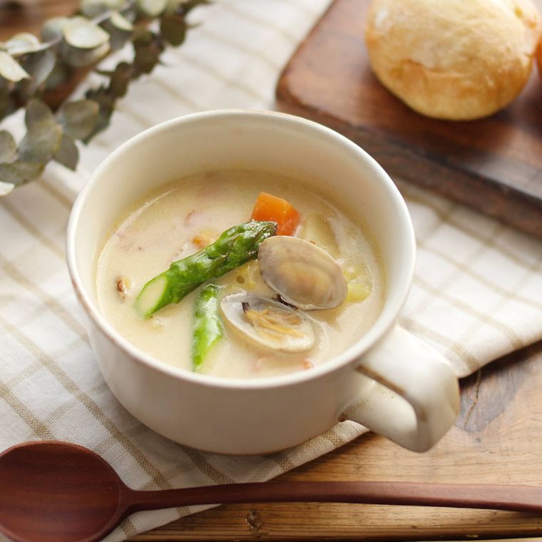 片手の白いマグカップに盛りつけられた春野菜とあさりのミルクみそスープ