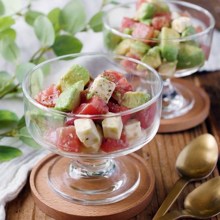 透明のグラスにアボカドとトマトのクリームチーズサラダが盛られ、木のコースターの上に置かれている