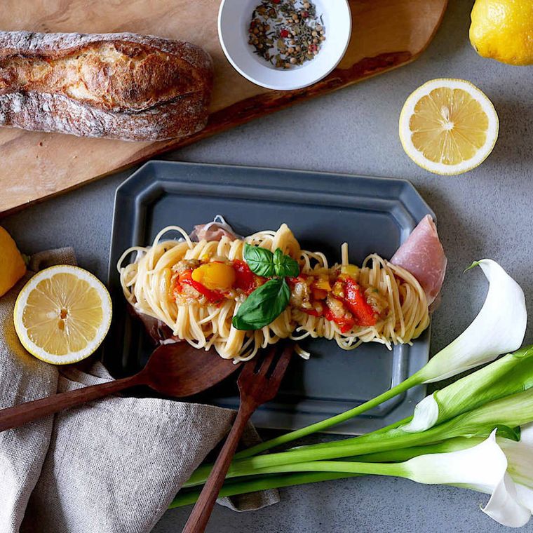 テーブルの上にある灰色の皿に盛られたペペロナータの冷製パスタや、数種類の食材と白い花