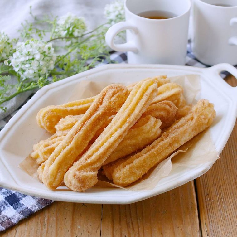 テーブルに白い皿に入ったチュロス、マグカップ、花が置かれている