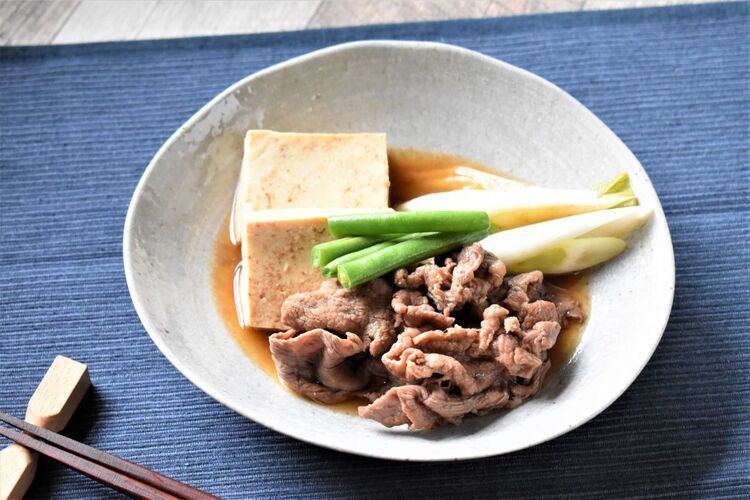 青いクロスの上、白い器に盛られた肉豆腐。牛肉、ねぎ、豆腐、いんげんが顔をのぞかせている