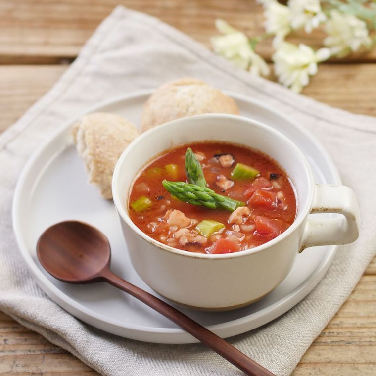 白いスープカップに野菜とタコのもち麦トマトスープが入っていてパンと木製スプーンが添えてある様子