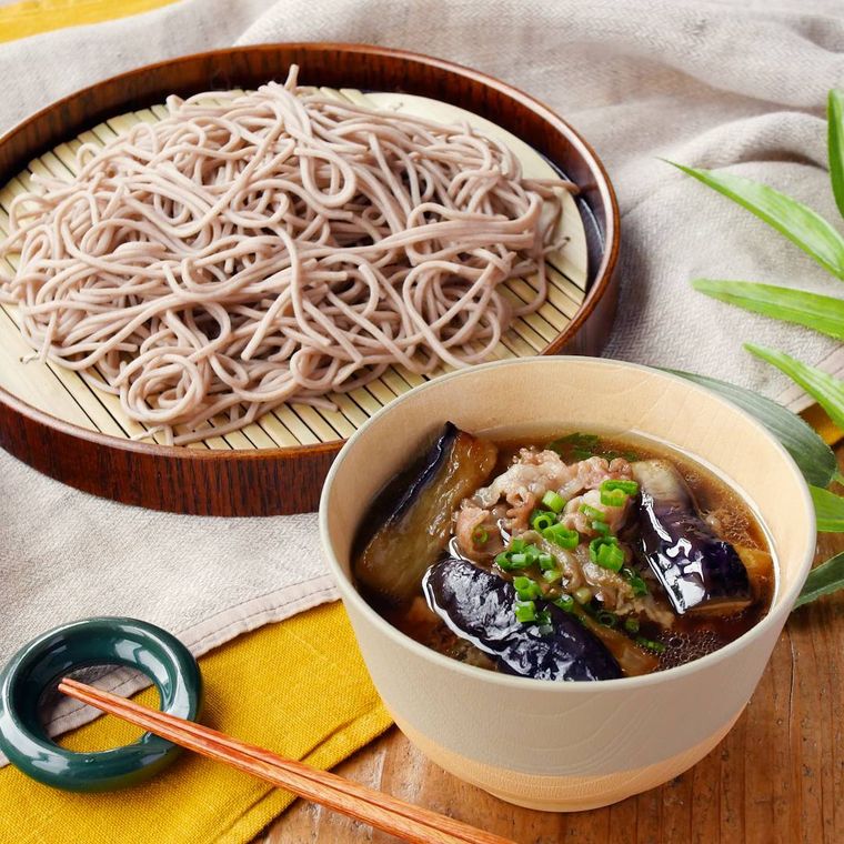灰色の汁椀に盛りつけられた豚バラなすのつけ汁と茶色の丸い皿に盛りつけられたそば
