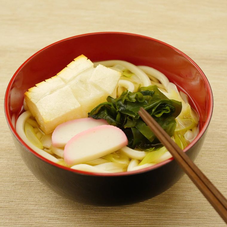 黒のお椀に入っている餅とかまぼことわかめがのってるうどん