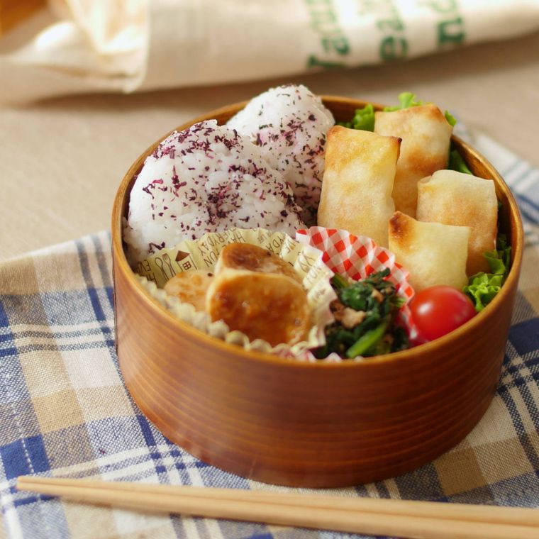 木の丸いお弁当箱におにぎりやミニトマト、ほうれん草の和え物、エリンギバター醤油、焼き春巻きが入っている