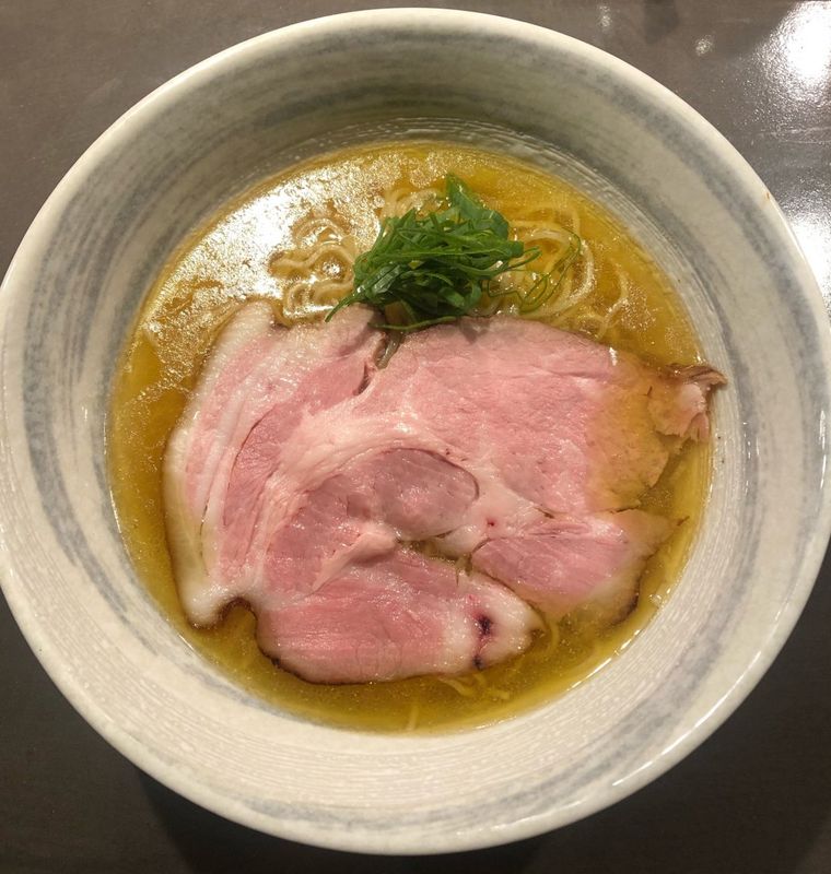 どんぶりの中にねぎ、大きいチャーシューがのった塩ラーメンが入って置いてある