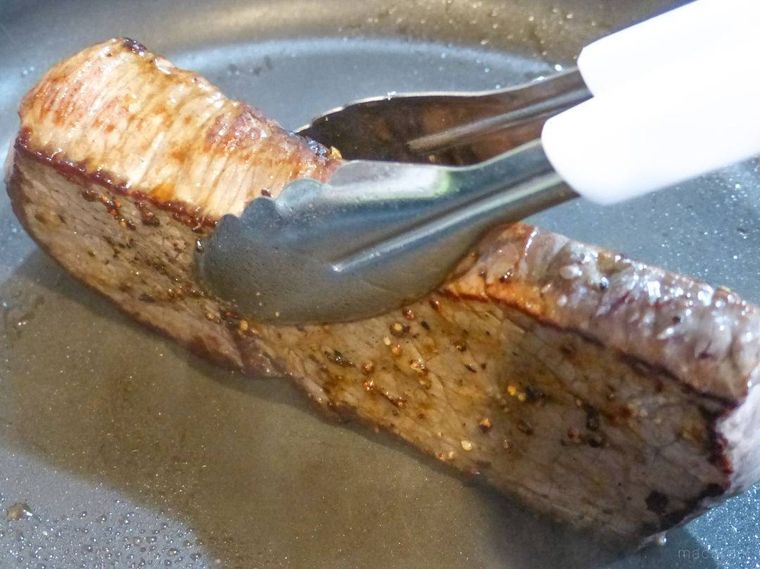 トングでフライパンに牛モモ肉の側面を押しつけて焼く様子