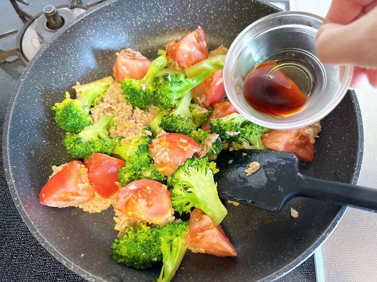 トマト、ツナ缶、ブロッコリーを炒めたところにしょうゆを加える様子